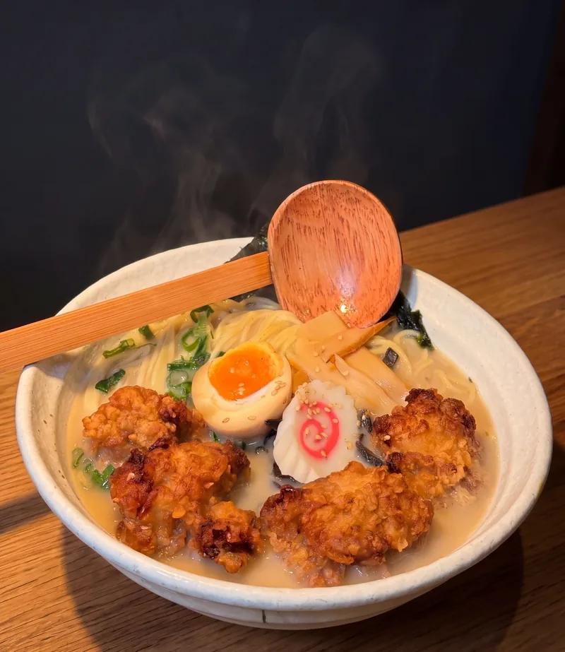 Karaage tonkotsu ramen with a jammy egg, narutomaki and a wooden ladle across the bowl, on timber
