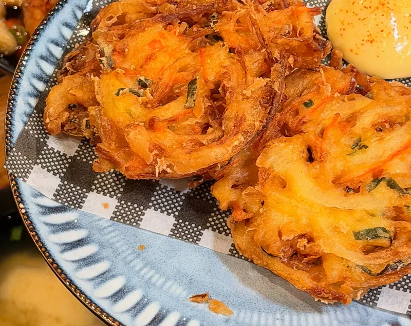 Two crisp kakiage fritters on a checked paper liner with spicy mayo